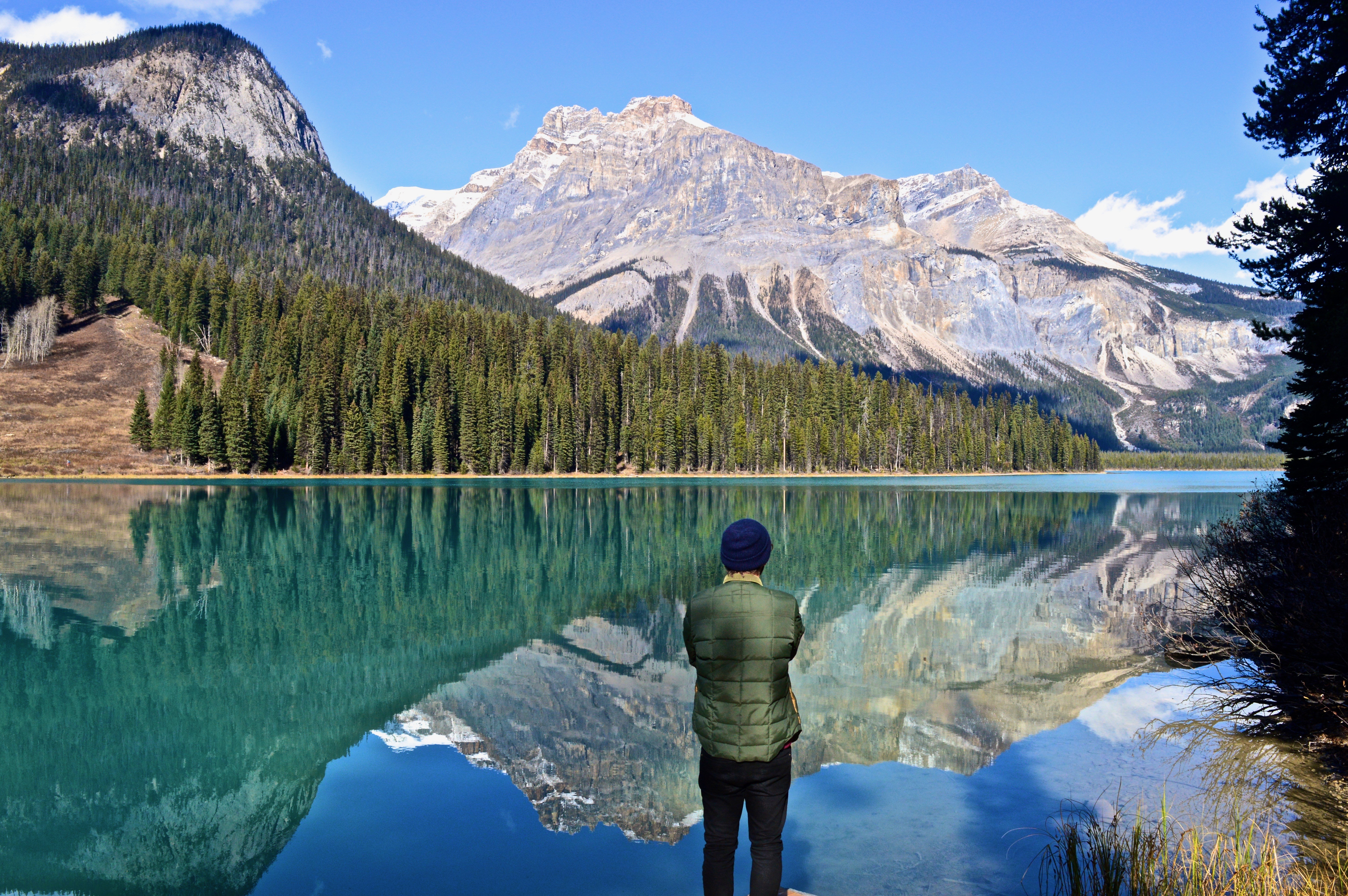 Een persoon die bij een turquoise meer in Canada staat en uitkijkt over de beboste kustlijn en de torenhoge toppen van de Rocky Mountains die weerspiegeld worden in het stille water.