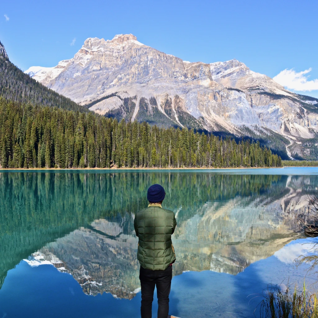 Een persoon die bij een turquoise meer in Canada staat en uitkijkt over de beboste kustlijn en de torenhoge toppen van de Rocky Mountains die weerspiegeld worden in het stille water.