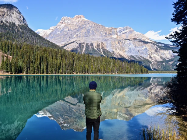 Een persoon die bij een turquoise meer in Canada staat en uitkijkt over de beboste kustlijn en de torenhoge toppen van de Rocky Mountains die weerspiegeld worden in het stille water.