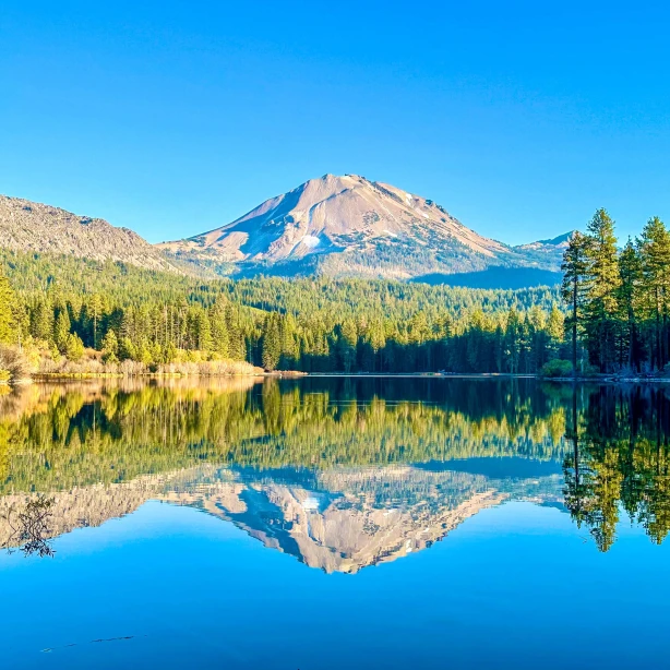 Een rustig meer in Californië dat de omringende dennenbossen weerspiegelt en de opvallende vulkanische piek die in de verte oprijst onder een helderblauwe hemel.