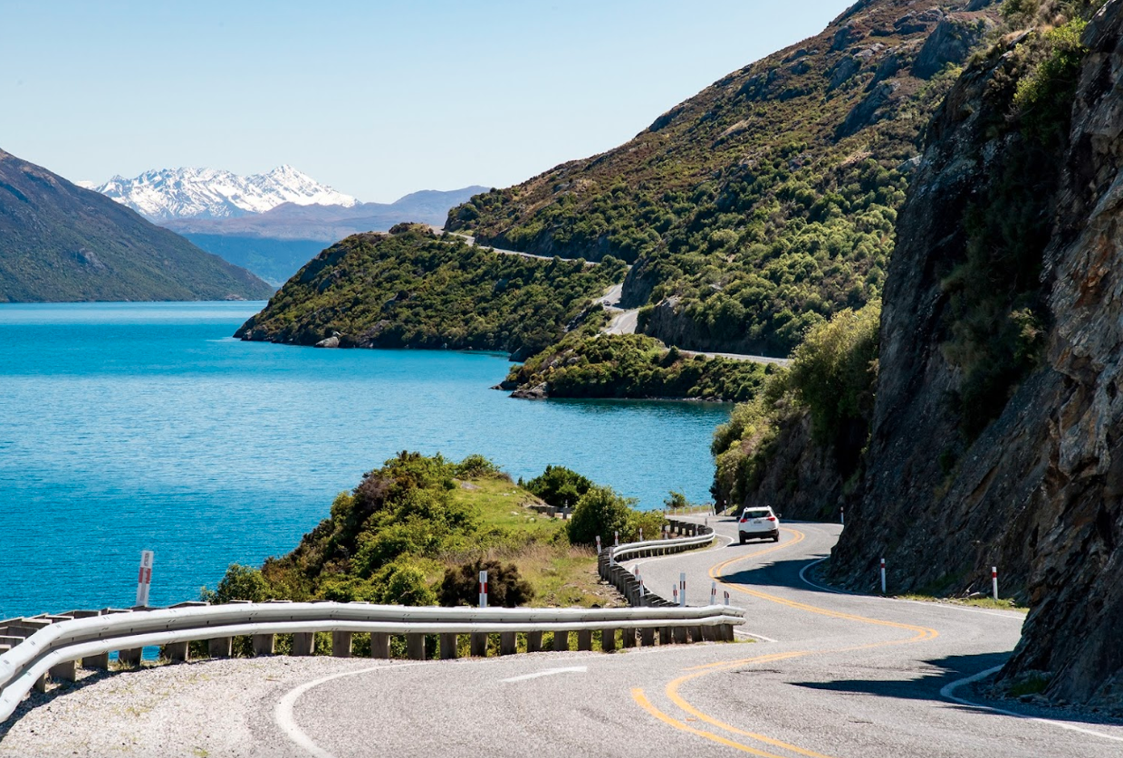 Een auto rijdt naast een blauw meer tussen de bergen in Nieuw-Zeeland