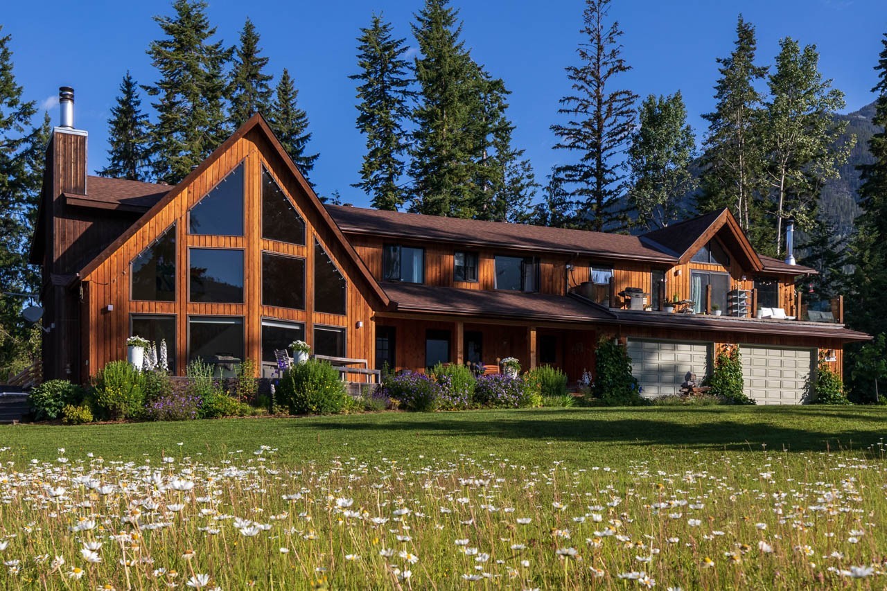 Grote houten lodge met kamerhoge ramen en een wilde bloemenweide op de voorgrond, omgeven door hoge sparren en bergtoppen in de buurt van Golden, British Columbia, Canada.