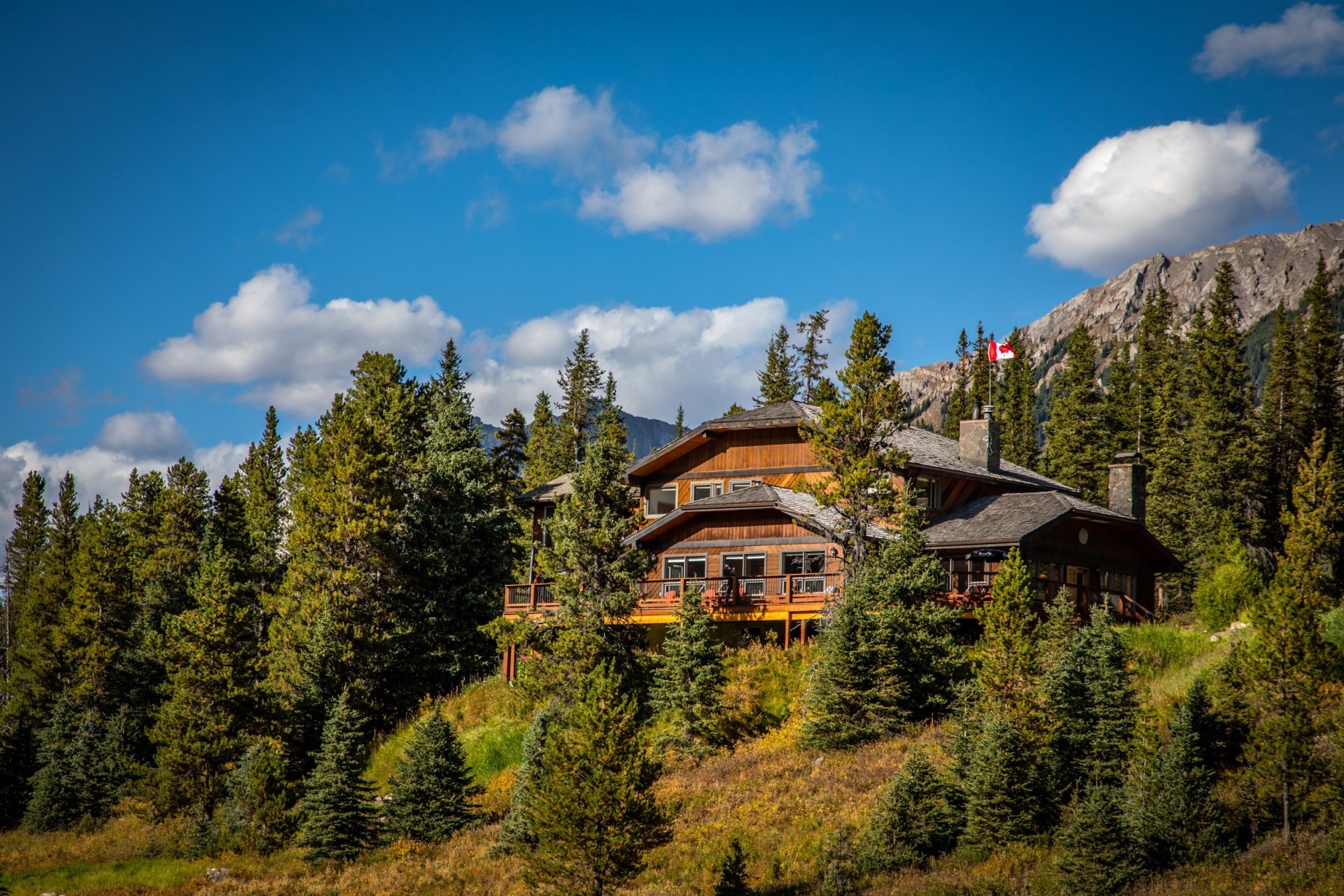 Houten berghut met rondomlopend terras, genesteld tussen sparrenbomen en herfstbladeren in Kananaskis Country, Alberta, Canada, met rotsachtige bergtoppen en een Canadese vlag op de achtergrond.