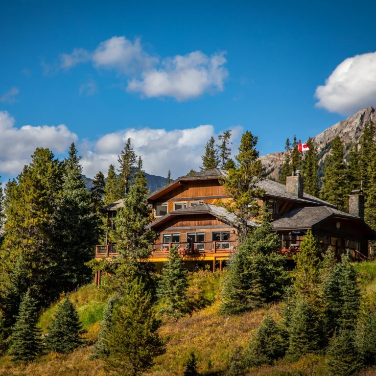 Houten berghut met rondomlopend terras, genesteld tussen sparrenbomen en herfstbladeren in Kananaskis Country, Alberta, Canada, met rotsachtige bergtoppen en een Canadese vlag op de achtergrond.