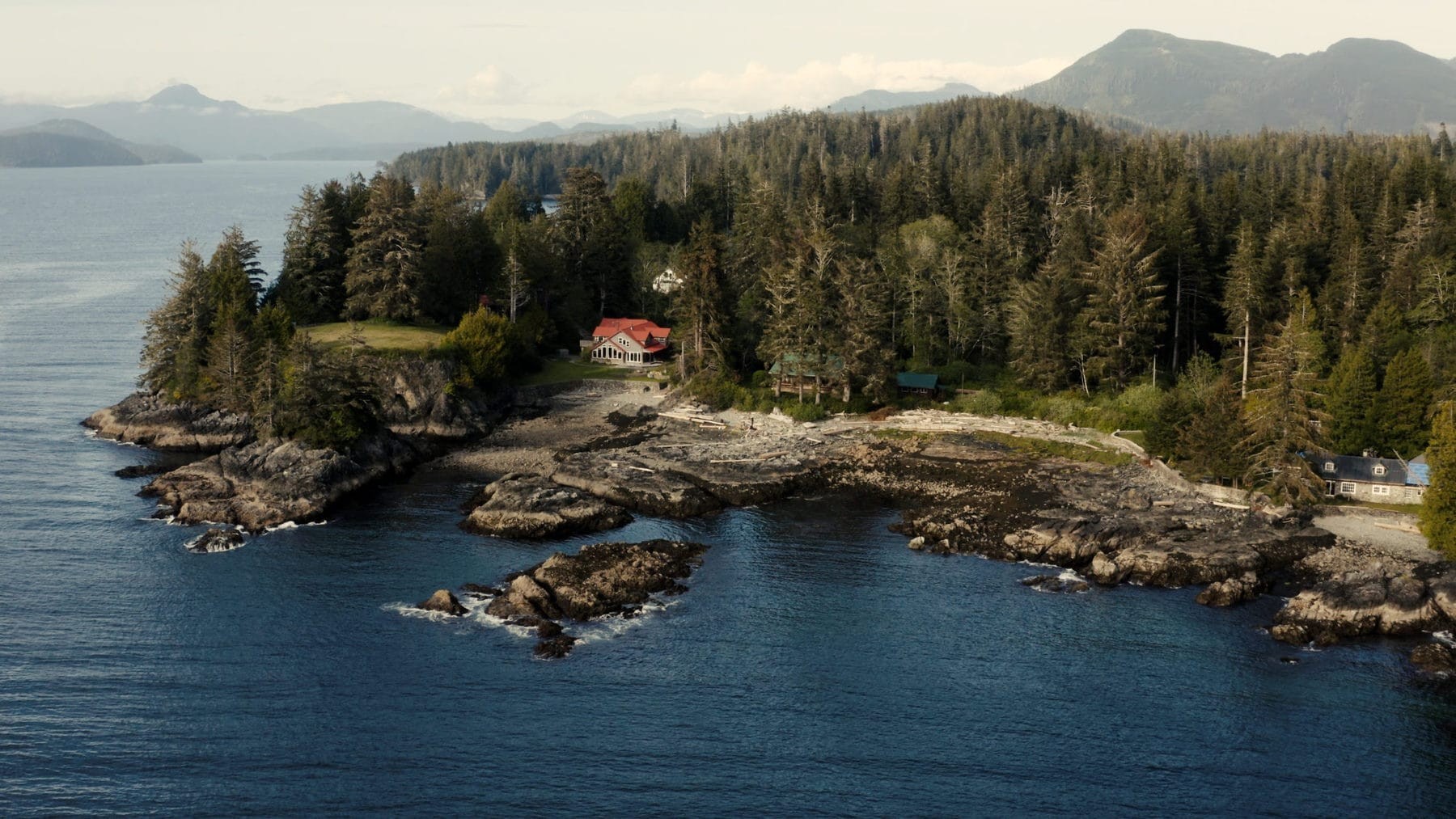 Luchtfoto van een afgelegen wildernislodge aan een rotsachtige Pacifische kustlijn in British Columbia, Canada, omgeven door dicht oerbos met bergketens zichtbaar aan de overkant van het water.