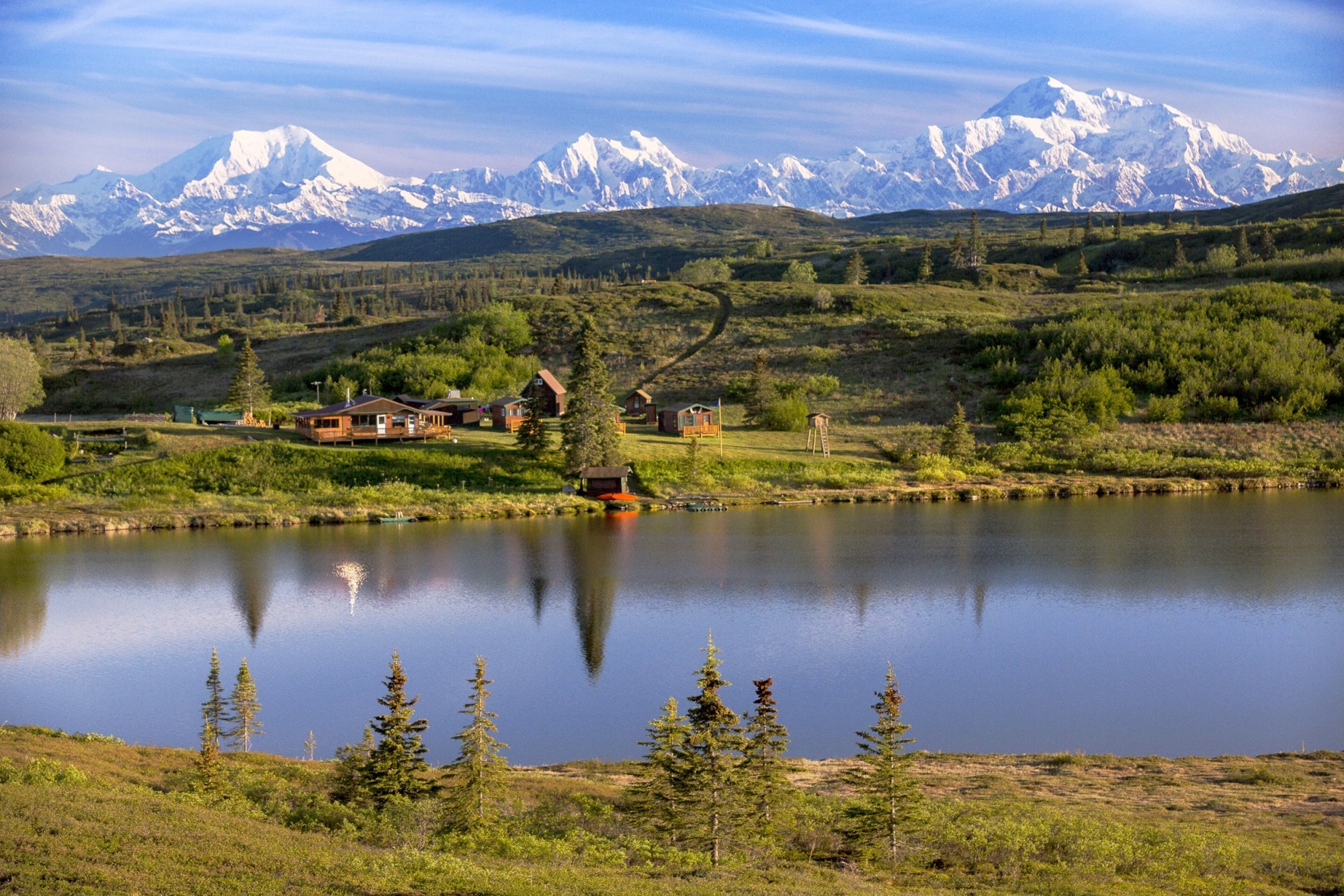 Afgelegen wildernislodge met houten hutten aan een rustig meer in Alaska, VS, met een adembenemend uitzicht op de met sneeuw bedekte toppen van de Denali-berg en het open toendralandschap.