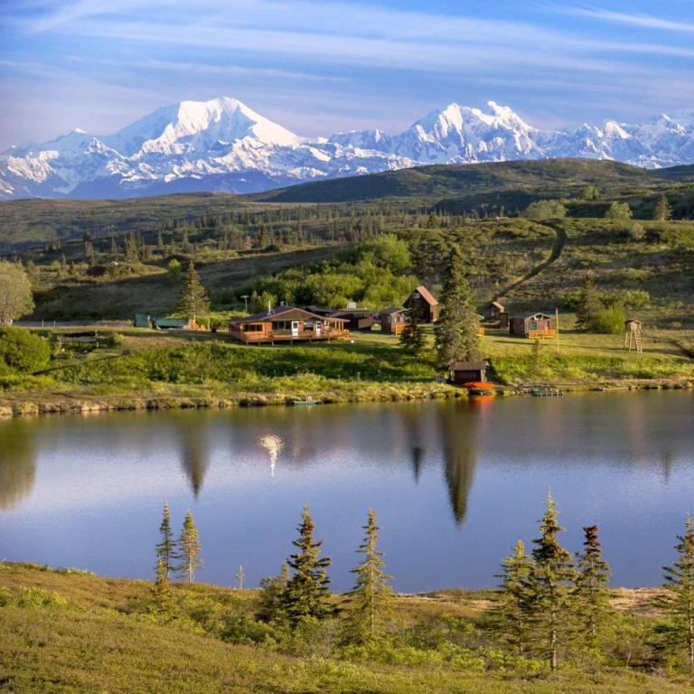 Afgelegen wildernislodge met houten hutten aan een rustig meer in Alaska, VS, met een adembenemend uitzicht op de met sneeuw bedekte toppen van de Denali-berg en het open toendralandschap.