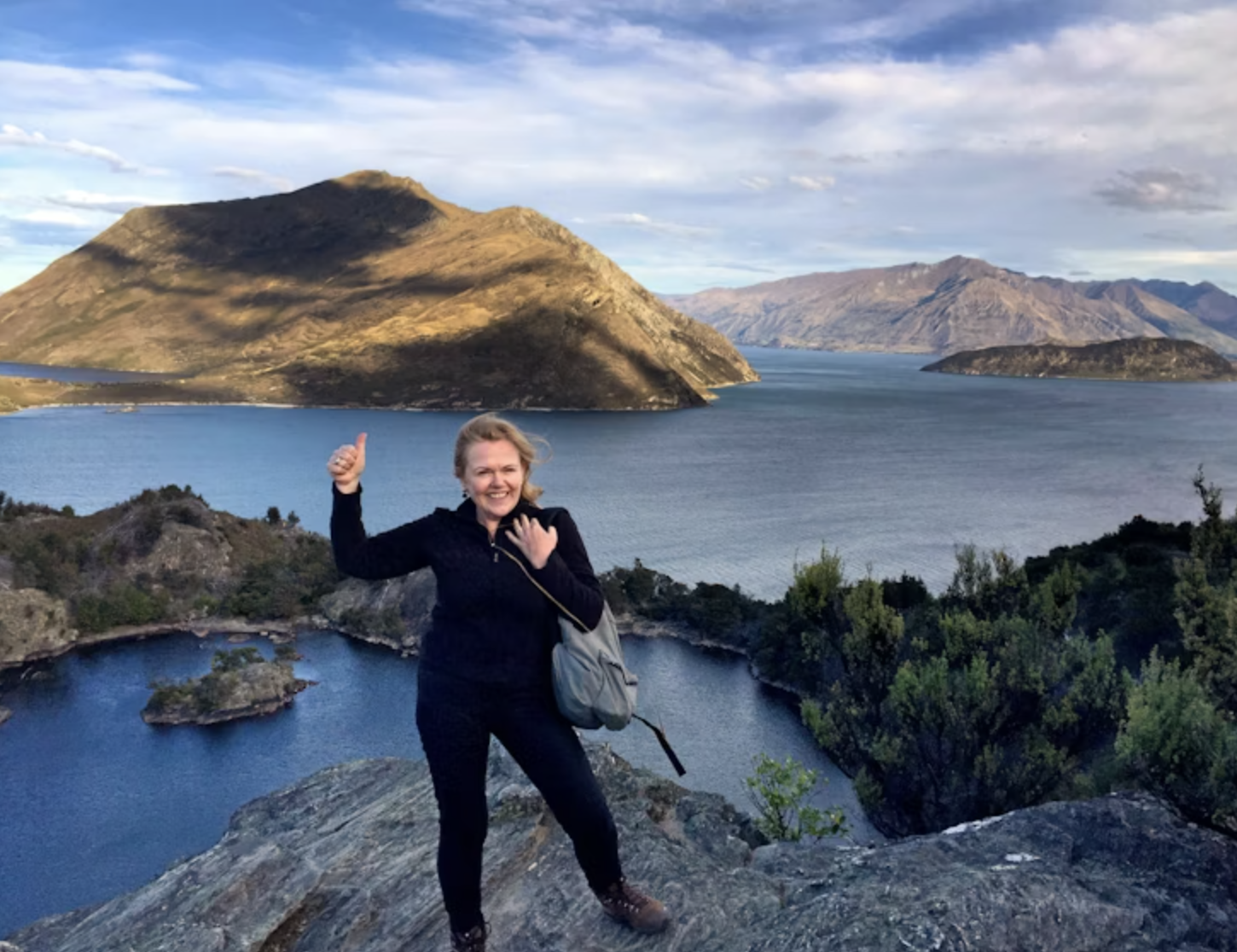 Gerrie glimlacht en geeft een duim omhoog naar de camera met de oceanen en bergen op de achtergrond