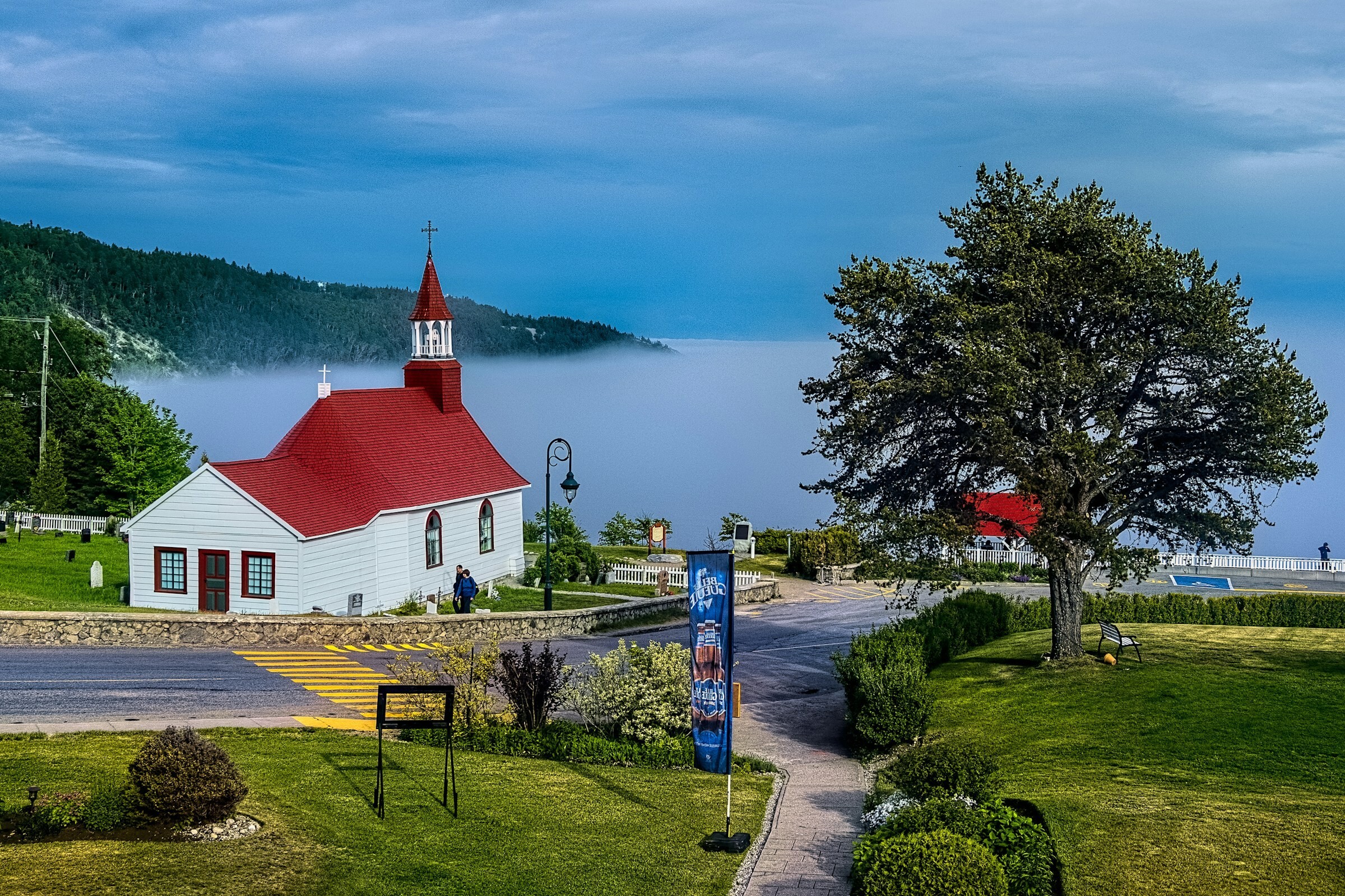 Een klein wit kerkje met een rood dak dat uitkijkt over een mistige baai in Canada, omringd door groene gazons en beboste heuvels.