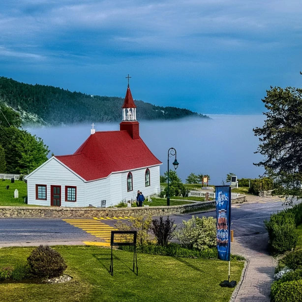 Een klein wit kerkje met een rood dak dat uitkijkt over een mistige baai in Canada, omringd door groene gazons en beboste heuvels.