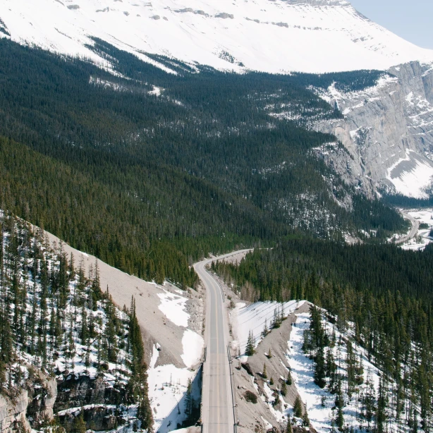 Een kronkelende bergweg in Canada die door dichte bossen leidt naar hoge, besneeuwde bergtoppen onder een heldere hemel.