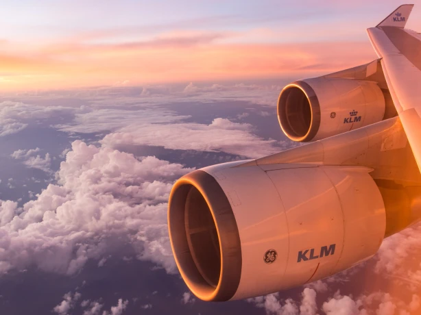 Een KLM vliegtuigvleugel en motoren gloeien in het zachte licht van een zonsopgang boven een laag wolken.