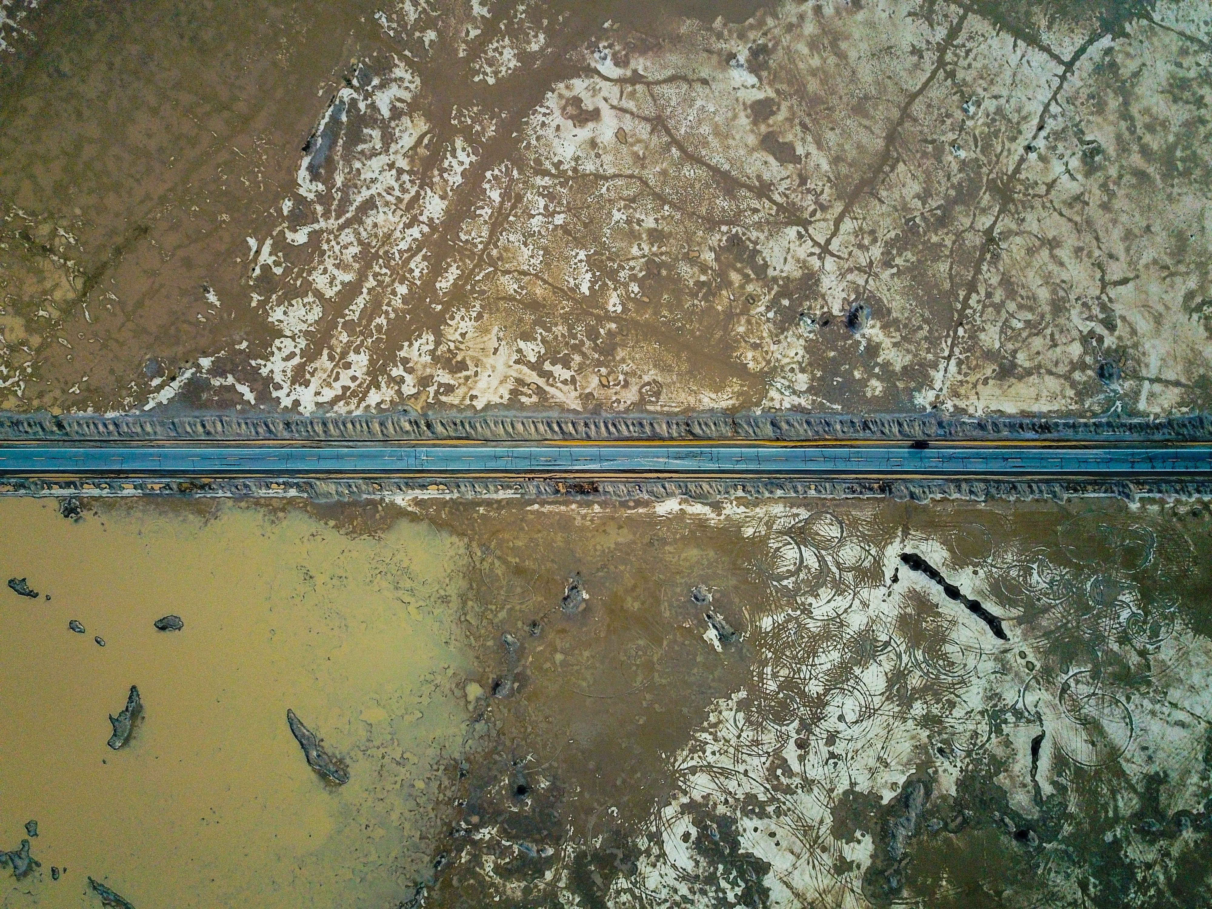 Een rechte weg dwars door de dorre woestijnvlakte van Death Valley, van bovenaf gezien met waterpartijen en gebarsten aarde aan weerszijden.