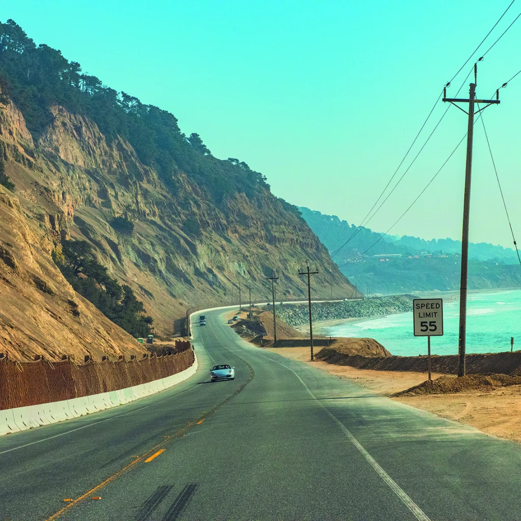 Een kustweg in Californië die buigt langs steile kliffen boven de oceaan, met auto's die langs het turquoise water rijden.