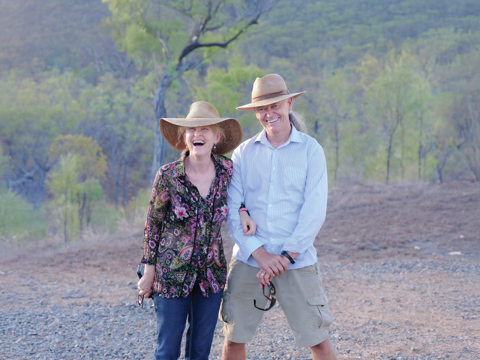 Glimlachende Australische gastheren met zonnehoeden, samen staand op een grindpad met bushland op de achtergrond.