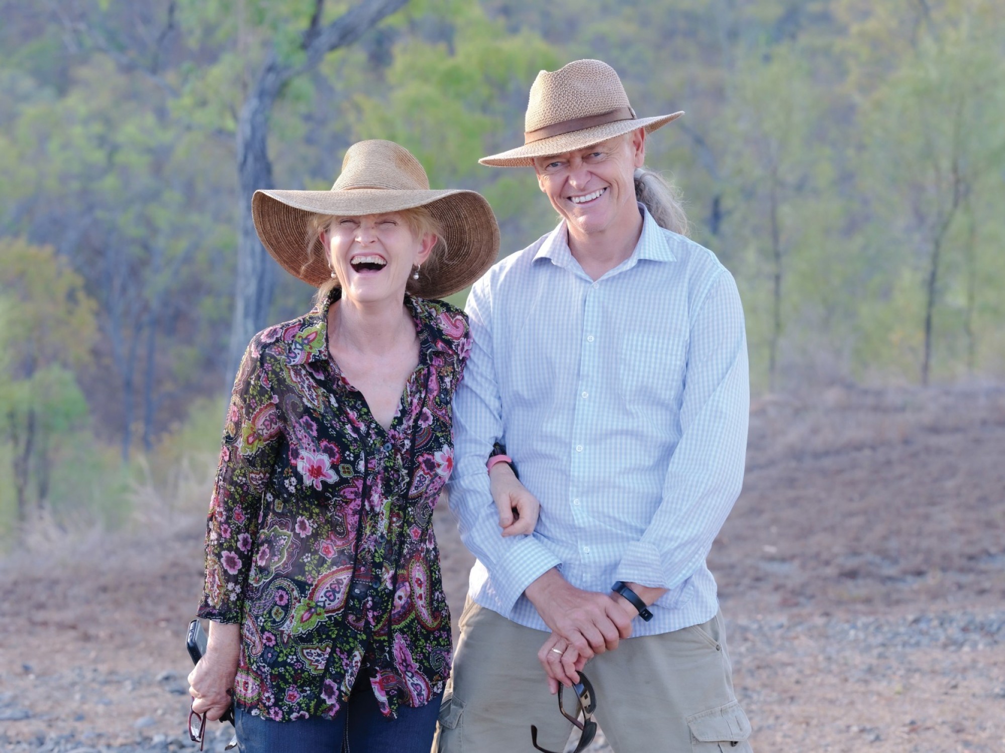 Lachend stel met zonnehoed, buiten staand op een grindpad met bushland op de achtergrond.