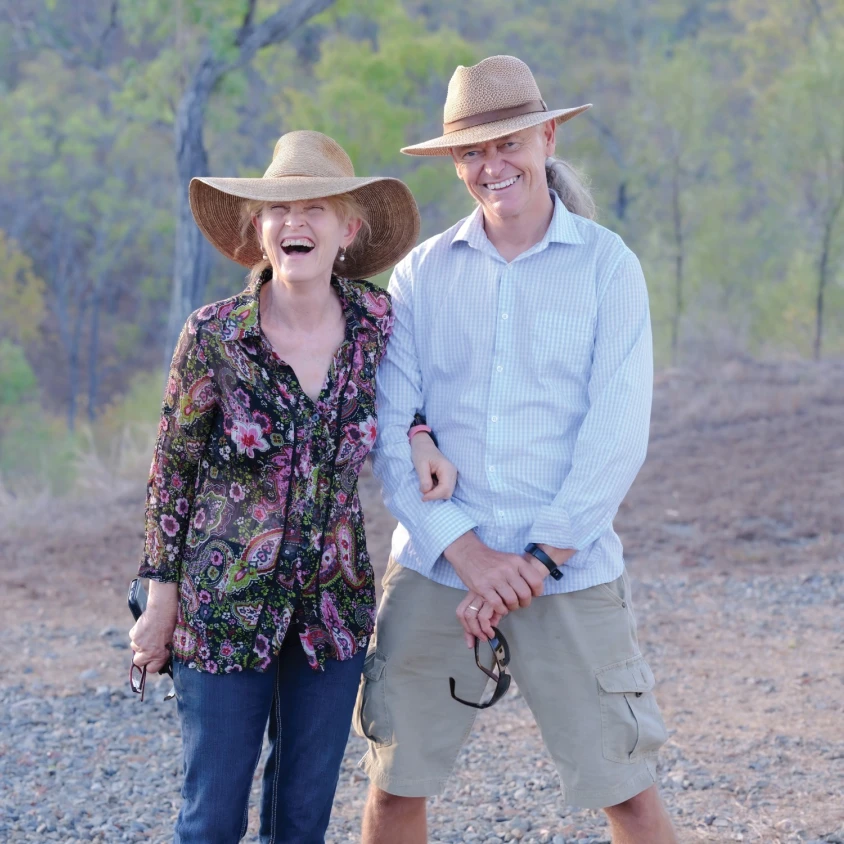 Lachend stel met zonnehoed, buiten staand op een grindpad met bushland op de achtergrond.