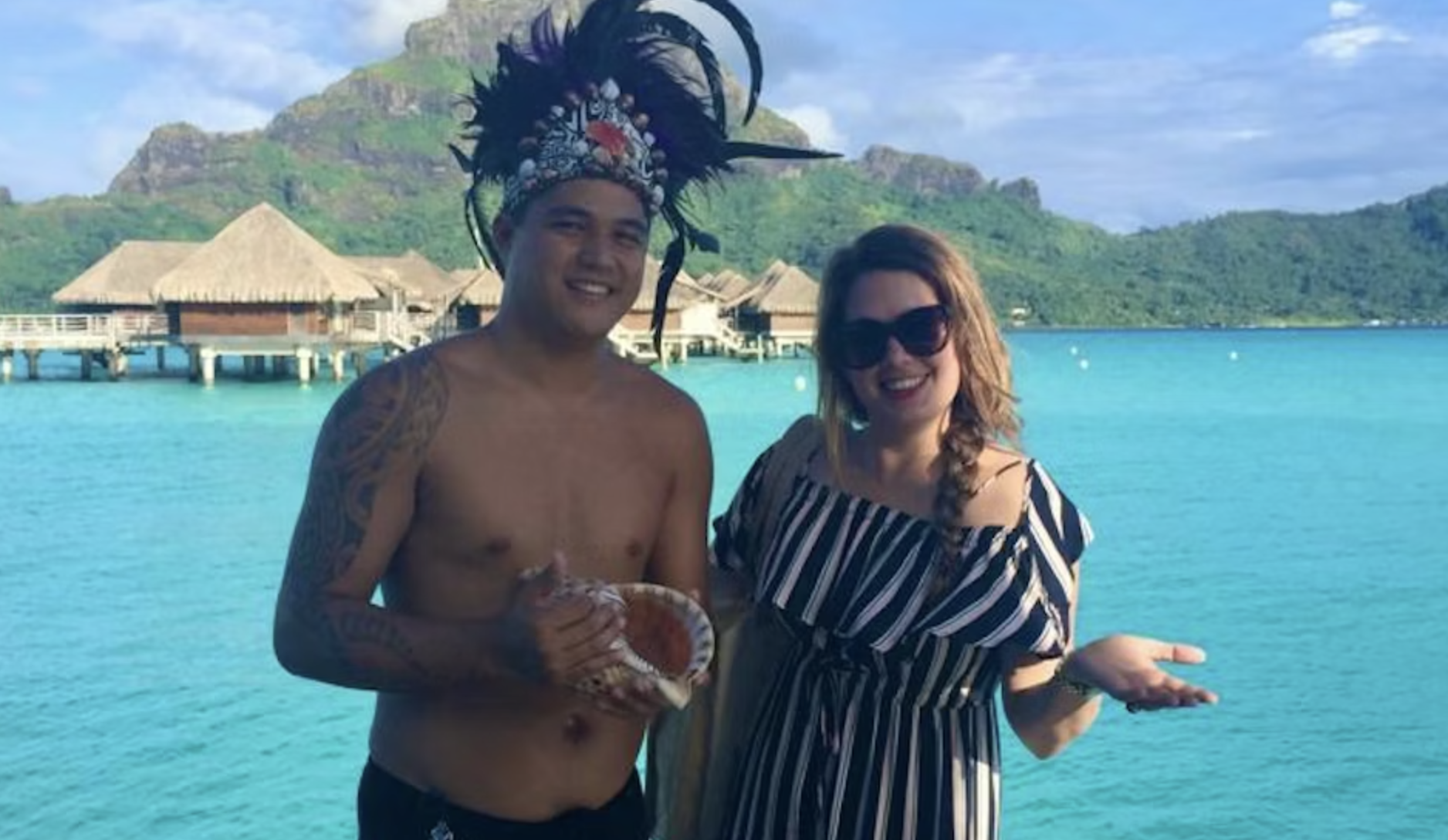 Sylvie en een Maori-man lachen naar de camera met de oceaan en bergen op de achtergrond