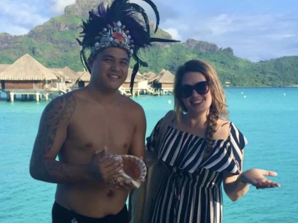 Sylvie en een Maori-man lachen naar de camera met de oceaan en bergen op de achtergrond