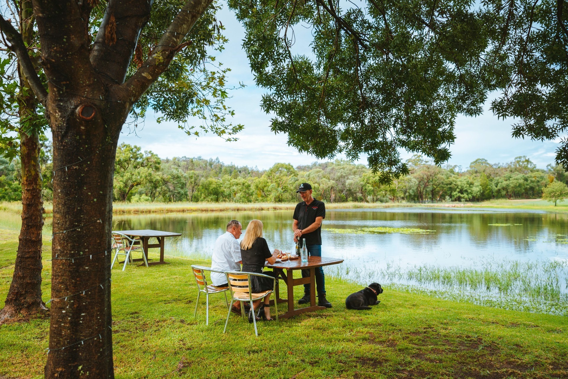 Echtpaar krijgt een wijnproeverij met gids, gezeten bij een meer met een zwarte hond in Australië