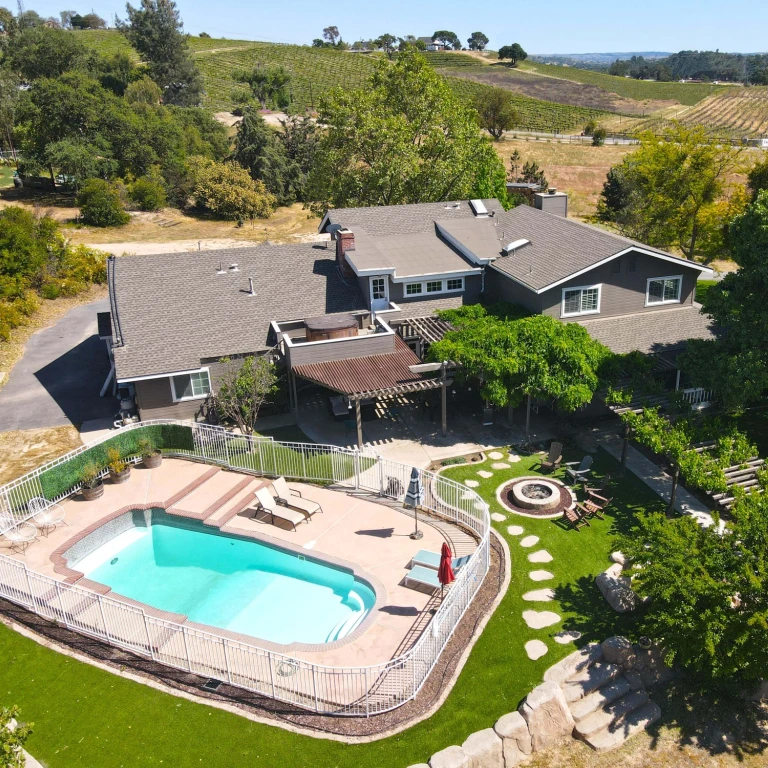 Luchtfoto van een vakantiehuis in een wijngaard in Centraal-Californië, VS, met een privézwembad, vuurplaats en pergola in aangelegde tuinen, omgeven door glooiende heuvels met wijngaarden.