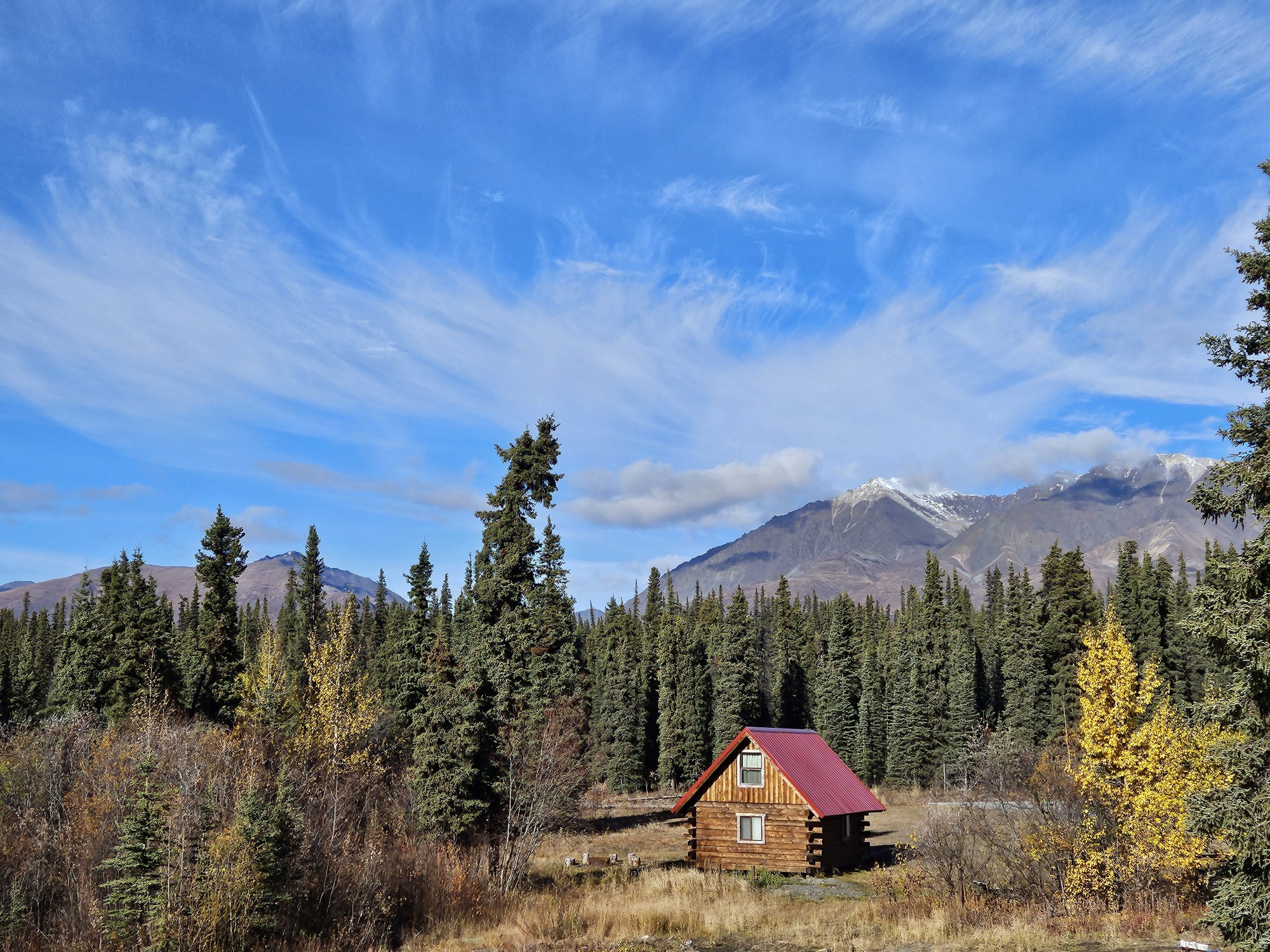Eenzame blokhut met een rood dak, genesteld tussen sparrenbomen en herfstbladeren in Wrangell-St. Elias National Park, Alaska, VS, met besneeuwde bergtoppen onder een dramatische blauwe lucht.