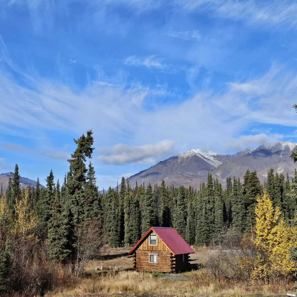 Eenzame blokhut met een rood dak, genesteld tussen sparrenbomen en herfstbladeren in Wrangell-St. Elias National Park, Alaska, VS, met besneeuwde bergtoppen onder een dramatische blauwe lucht.