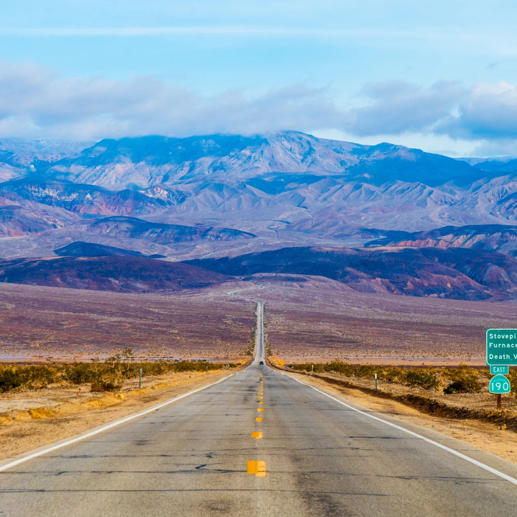 Een lange rechte weg die zich uitstrekt door de Mojavewoestijn van Californië naar de bergen van Death Valley, met woestijnvlakten en bergkammen in de verte onder een heldere hemel.