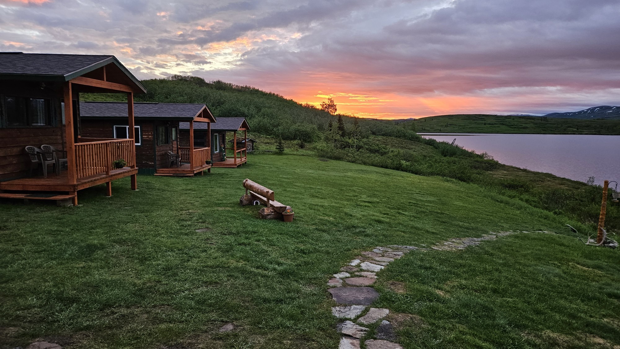 Wildernislodges met overdekte veranda's op een groene heuvel met uitzicht op een meer in Alaska, VS, onder een dramatische vuurrode en oranje middernachtelijke zonsonderganghemel.