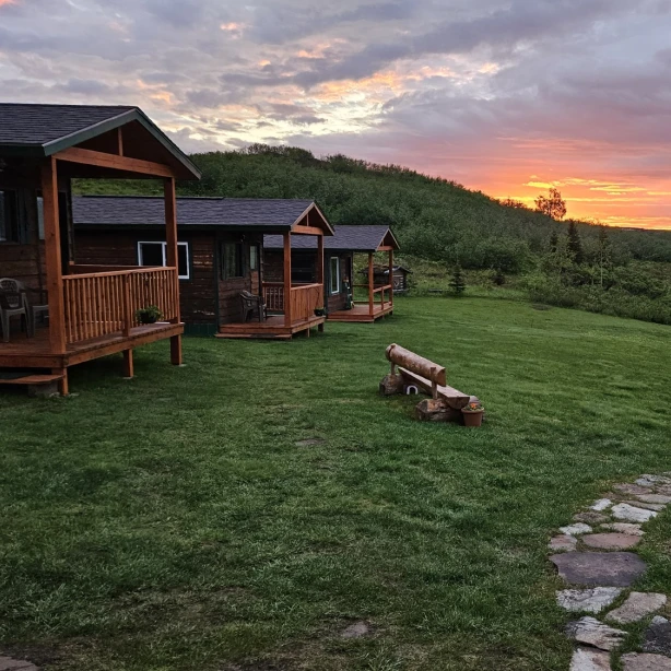 Wildernislodges met overdekte veranda's op een groene heuvel met uitzicht op een meer in Alaska, VS, onder een dramatische vuurrode en oranje middernachtelijke zonsonderganghemel.