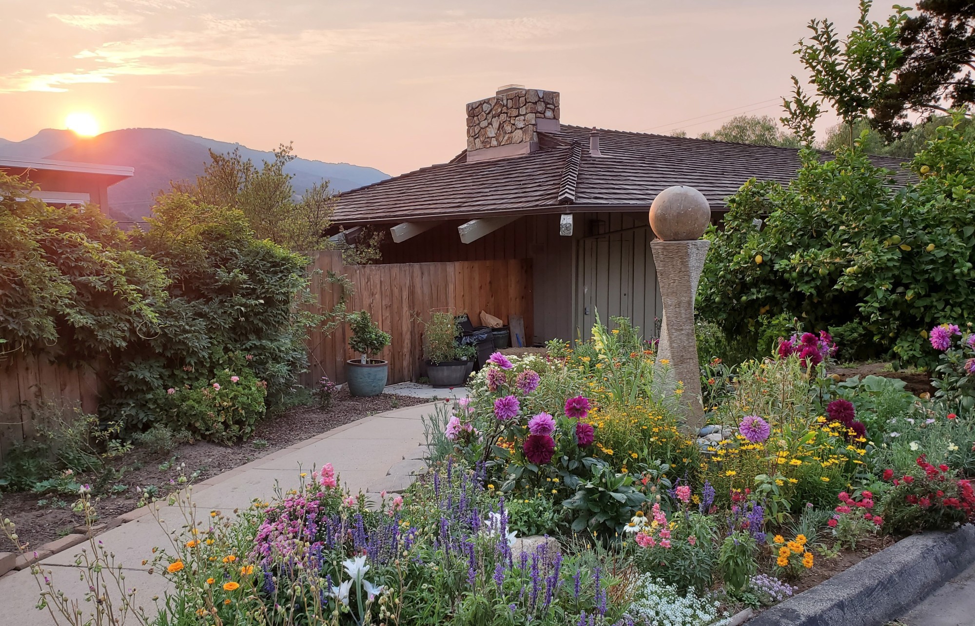 Charmante lodge cottage met een stenen schoorsteen, omgeven door een kleurrijke wilde bloementuin in Californië, VS, met glooiende heuvels en een warme zonsondergang aan de horizon.
