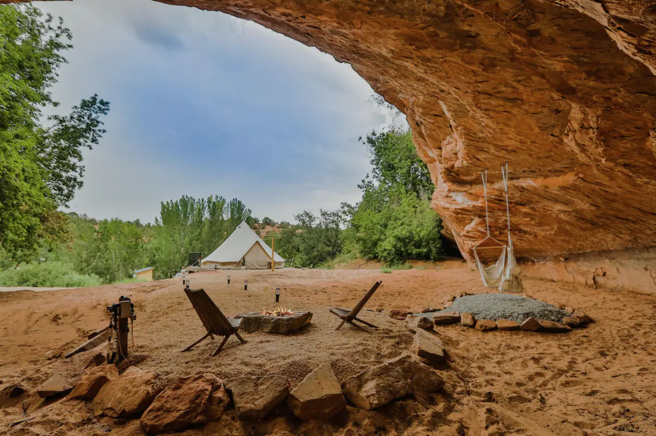 Unieke glampinglocatie onder een natuurlijke zandstenen rotsoverhang in Utah, VS, met een canvas bell-tent, kampvuur en houten stoelen in een rood woestijnlandschap.