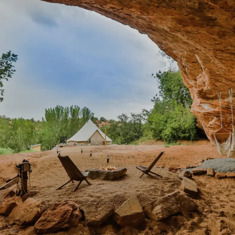 Unieke glampinglocatie onder een natuurlijke zandstenen rotsoverhang in Utah, VS, met een canvas bell-tent, kampvuur en houten stoelen in een rood woestijnlandschap.