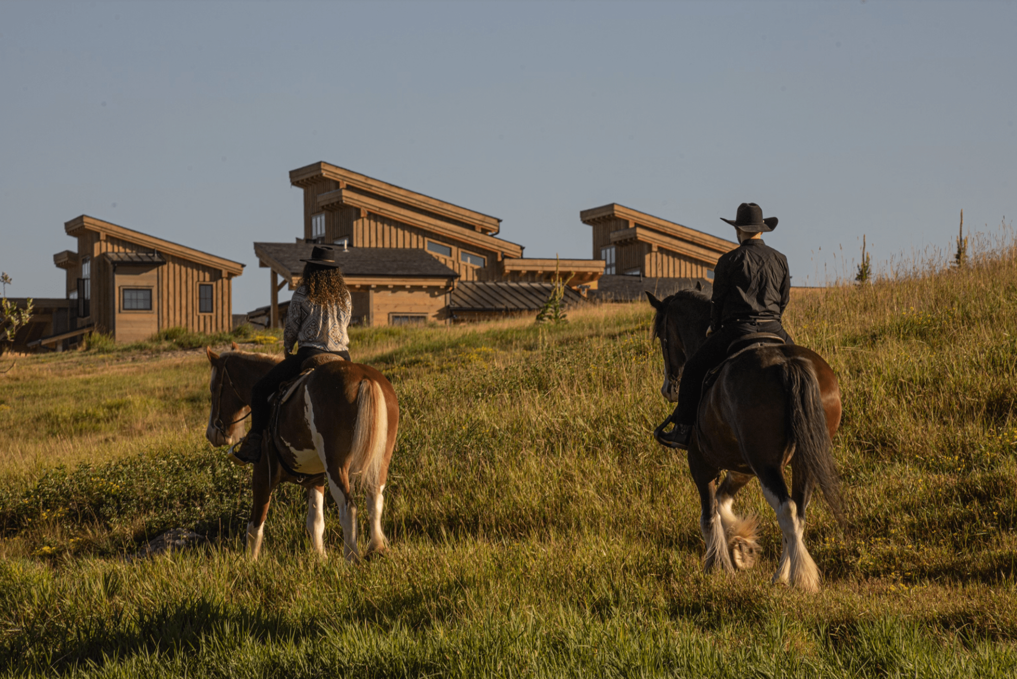 Twee ruiters met cowboyhoeden te paard die een met gras begroeide heuvel oversteken in de richting van een rustieke houten lodge in de buurt van Glacier National Park, Montana, VS, in het warme gouden licht van de middag.