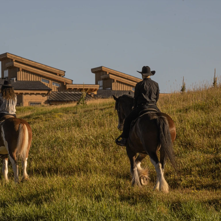Twee ruiters met cowboyhoeden te paard die een met gras begroeide heuvel oversteken in de richting van een rustieke houten lodge in de buurt van Glacier National Park, Montana, VS, in het warme gouden licht van de middag.