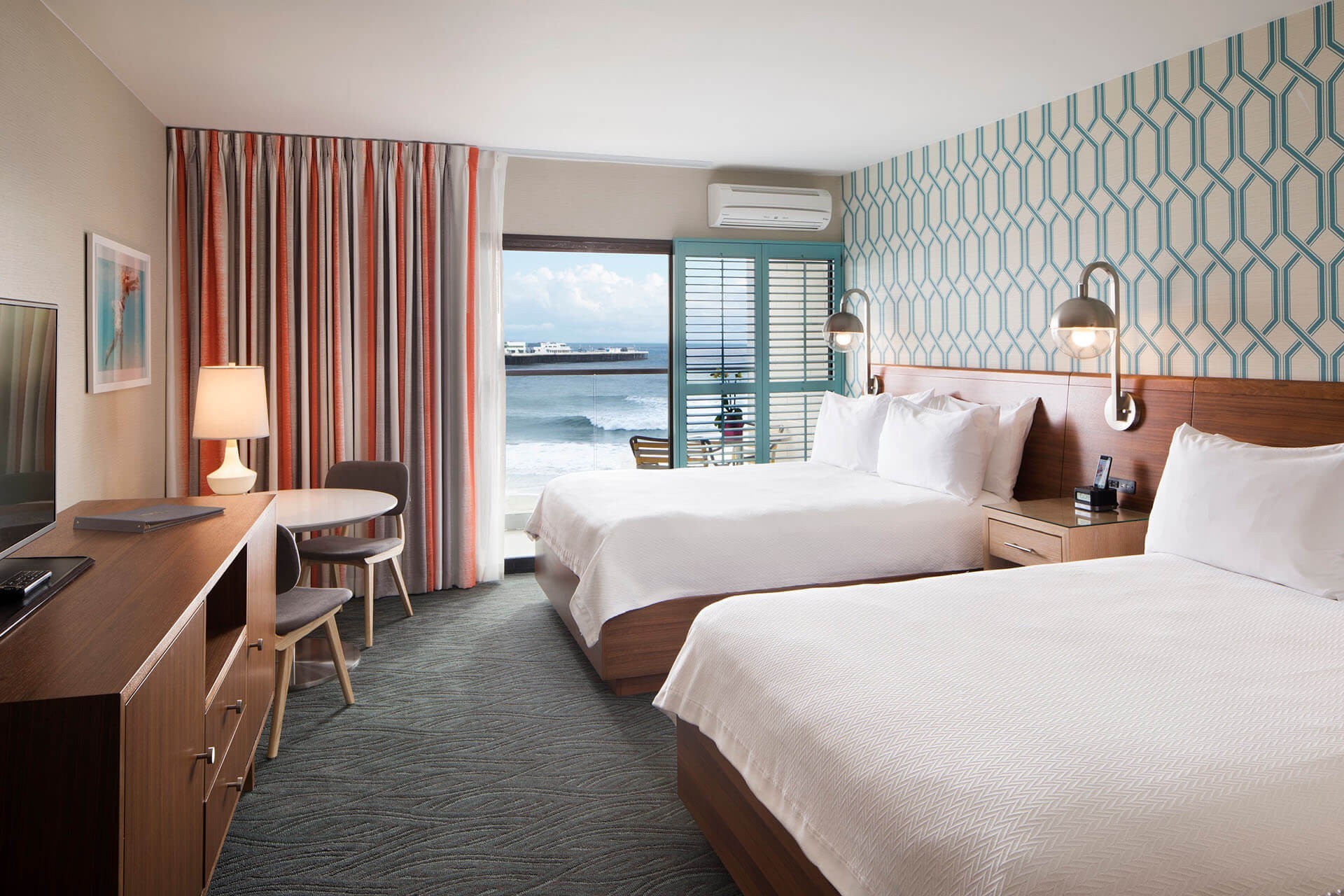 Lichte hotelkamer in retrostijl in Santa Cruz, Californië, VS, met twee queensize bedden, blauwgroen geometrisch behang en een balkon met uitzicht op de oceaan en de pier.