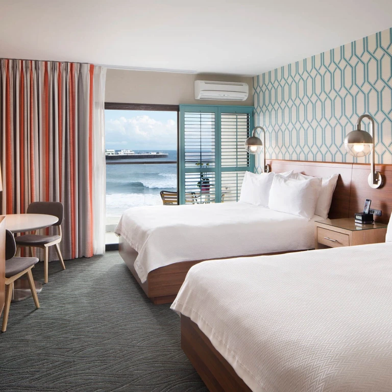 Lichte hotelkamer in retrostijl in Santa Cruz, Californië, VS, met twee queensize bedden, blauwgroen geometrisch behang en een balkon met uitzicht op de oceaan en de pier.