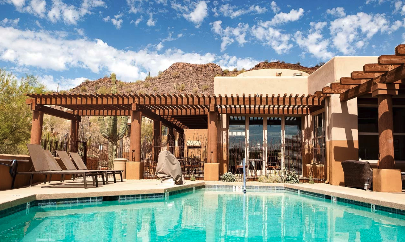 Resortzwembadterras in zuidwestelijke stijl in Scottsdale, Arizona, VS, met ligstoelen en een houten pergola tegen een achtergrond van rotsachtige woestijnheuvels en saguaro-cactussen onder een blauwe lucht.