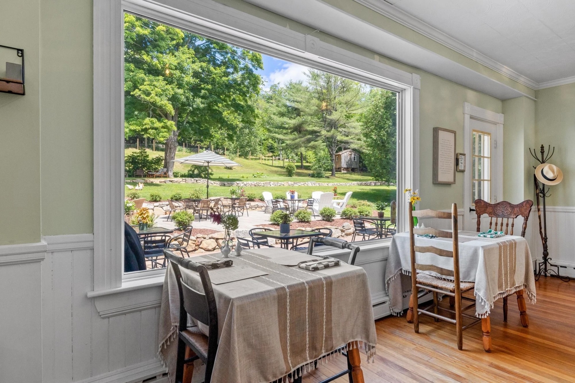 Gezellige bed & breakfast-eetkamer in de VS met met linnen gedekte tafels en een groot raam met uitzicht op een zonnig tuinterras en een groene, beboste heuvel.