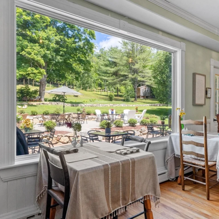 Gezellige bed & breakfast-eetkamer in de VS met met linnen gedekte tafels en een groot raam met uitzicht op een zonnig tuinterras en een groene, beboste heuvel.