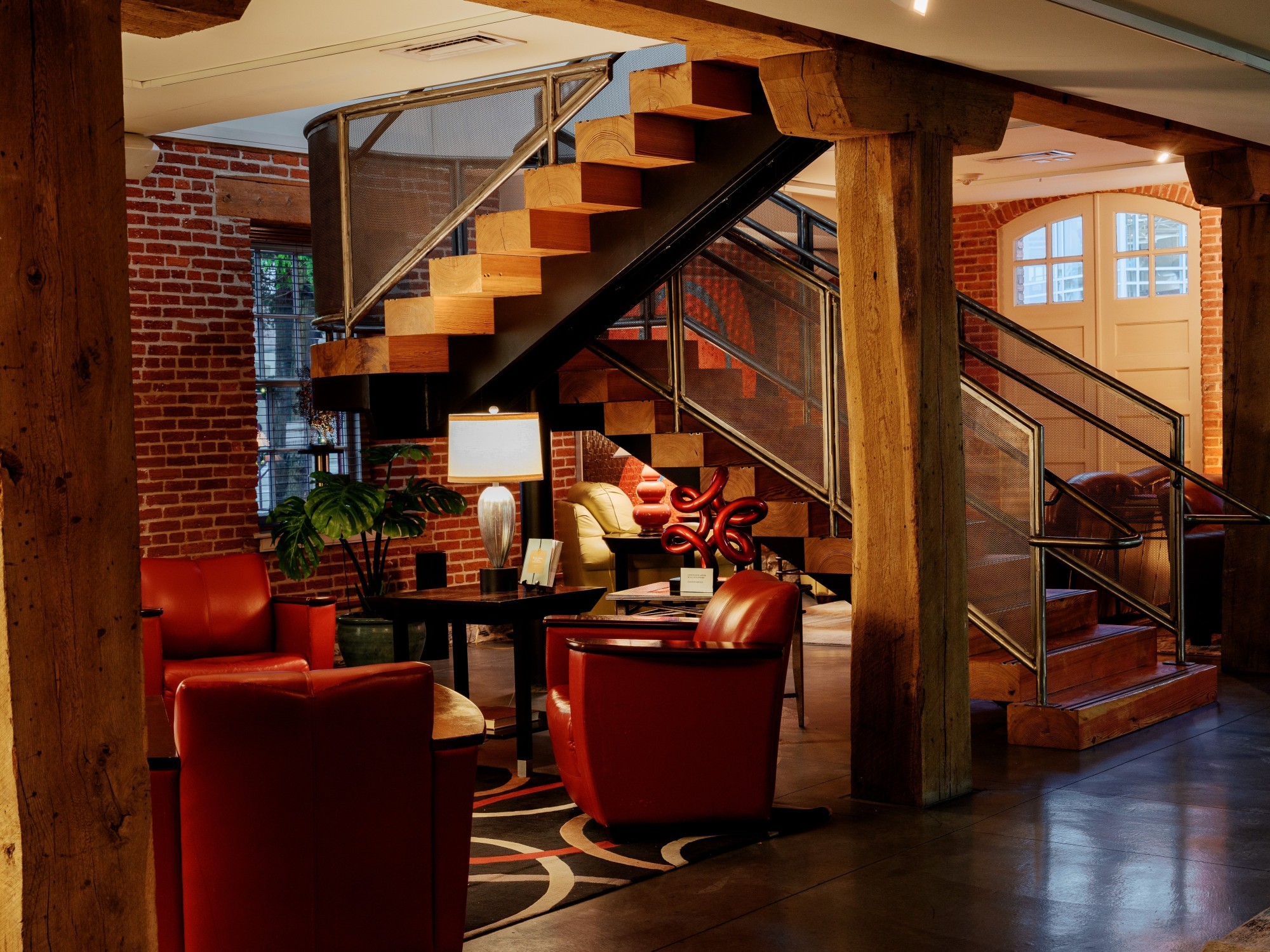 Stijlvolle lobby van een boetiekhotel in Pennsylvania, VS, met bakstenen muren, zware houten balken, rode leren fauteuils en een sculpturale zwevende trap in een gerenoveerd historisch gebouw.