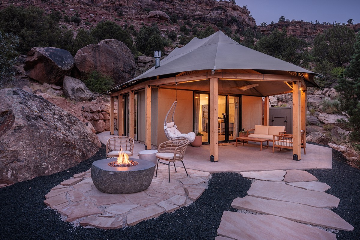 Luxe glamping-tent met een eigen stenen terras en vuurplaats bij zonsondergang, genesteld tussen rode rotsblokken in de buurt van Zion National Park, Utah, VS, met een warm, sfeervol interieur en een hangstoel.