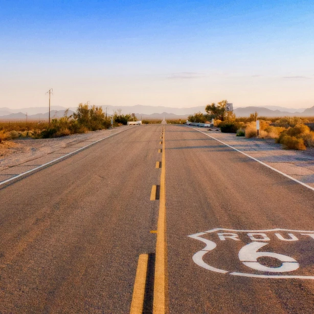 Een leeg stuk Route 66 door de woestijn bij zonsopgang, met lange schaduwen en bergsilhouetten in de verte.